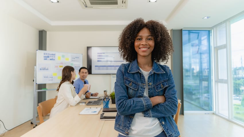 Young female Level 3 Financial Services Administrator apprentice standing in front of a table of with brainstorming colleagues at a meeting n a small office with a laptop and financial graphs on the table