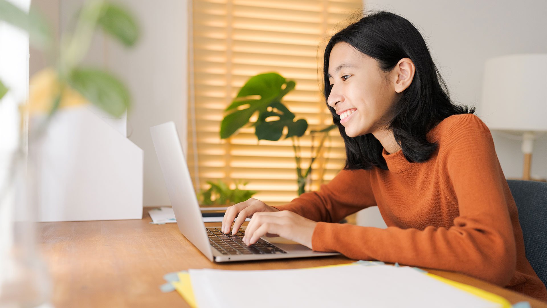 A young girl sitting at her desk, typing on her laptop and smiling.