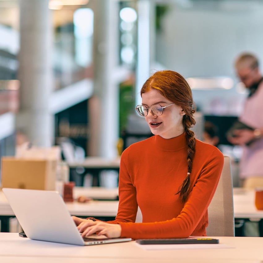 A young woman wearing glasses is sitting at a desk and typing on her laptop.