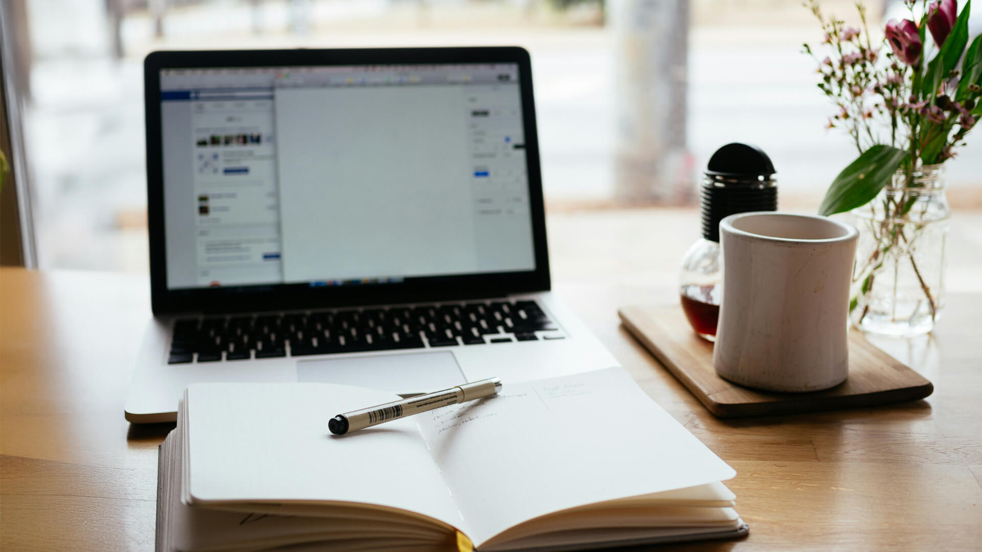 A laptop on a desk showing a document, with a notebook, pen and plant nearby.