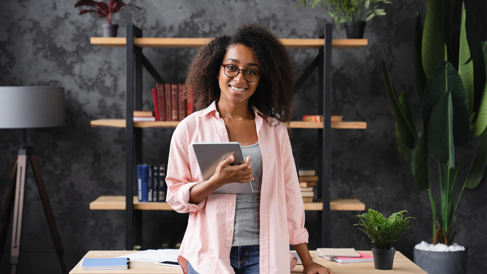 Student standing by a desk with a tablet, preparing to contact student services about a master’s course January start.