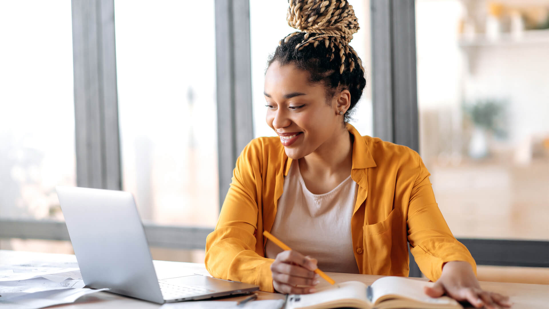 An online degree student sitting at a desk, smiling while working on a laptop and taking notes.