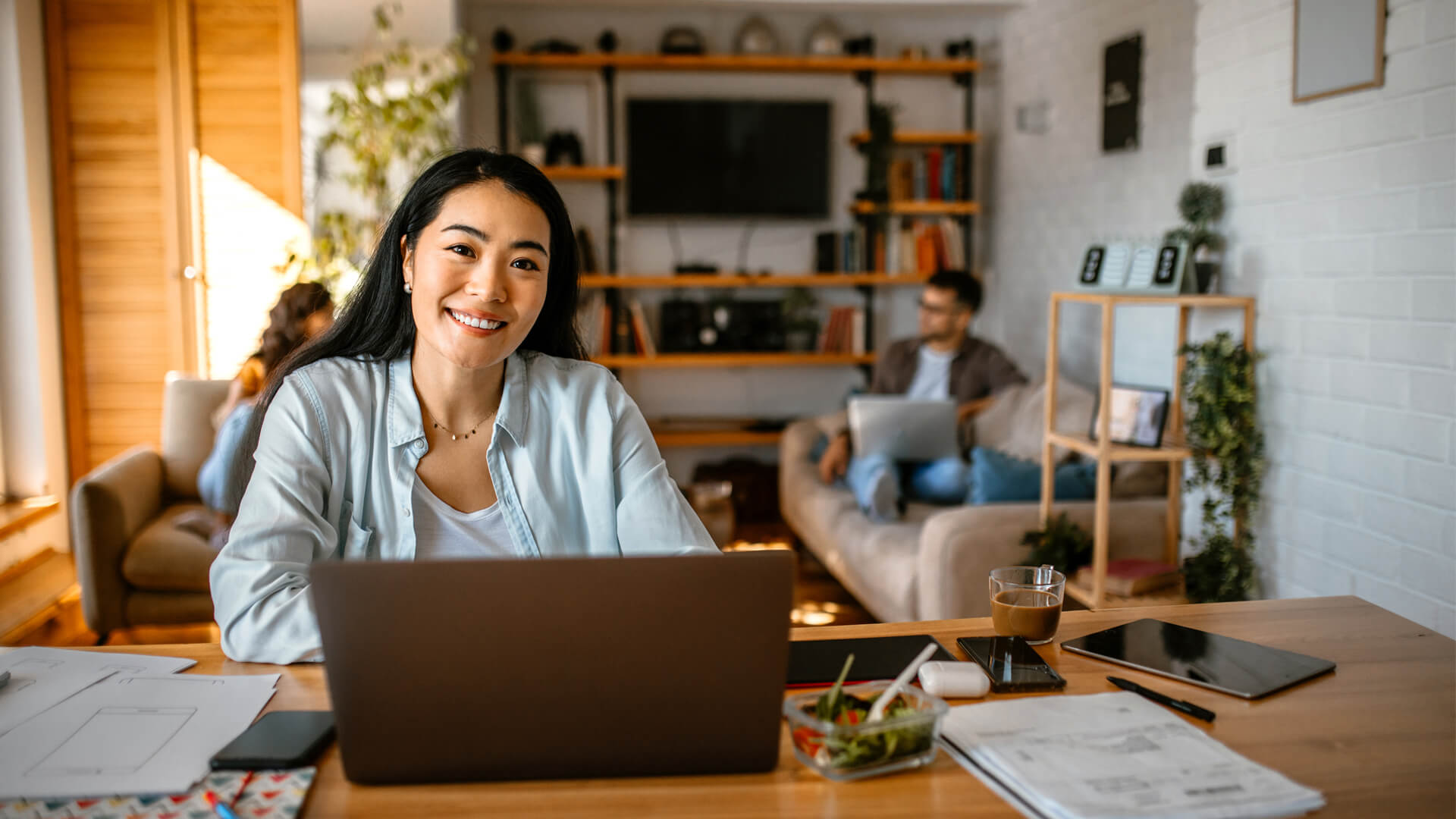 An online degree student sitting at a desk at home, looking at a laptop while their family sits in the background.