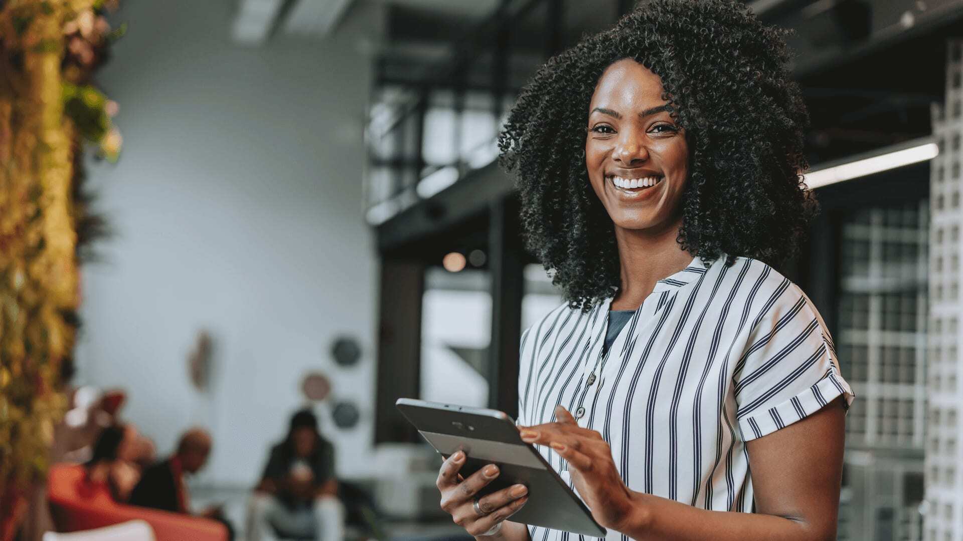 A woman in striped shirt smiling while holding a tablet in a busy indoor setting.