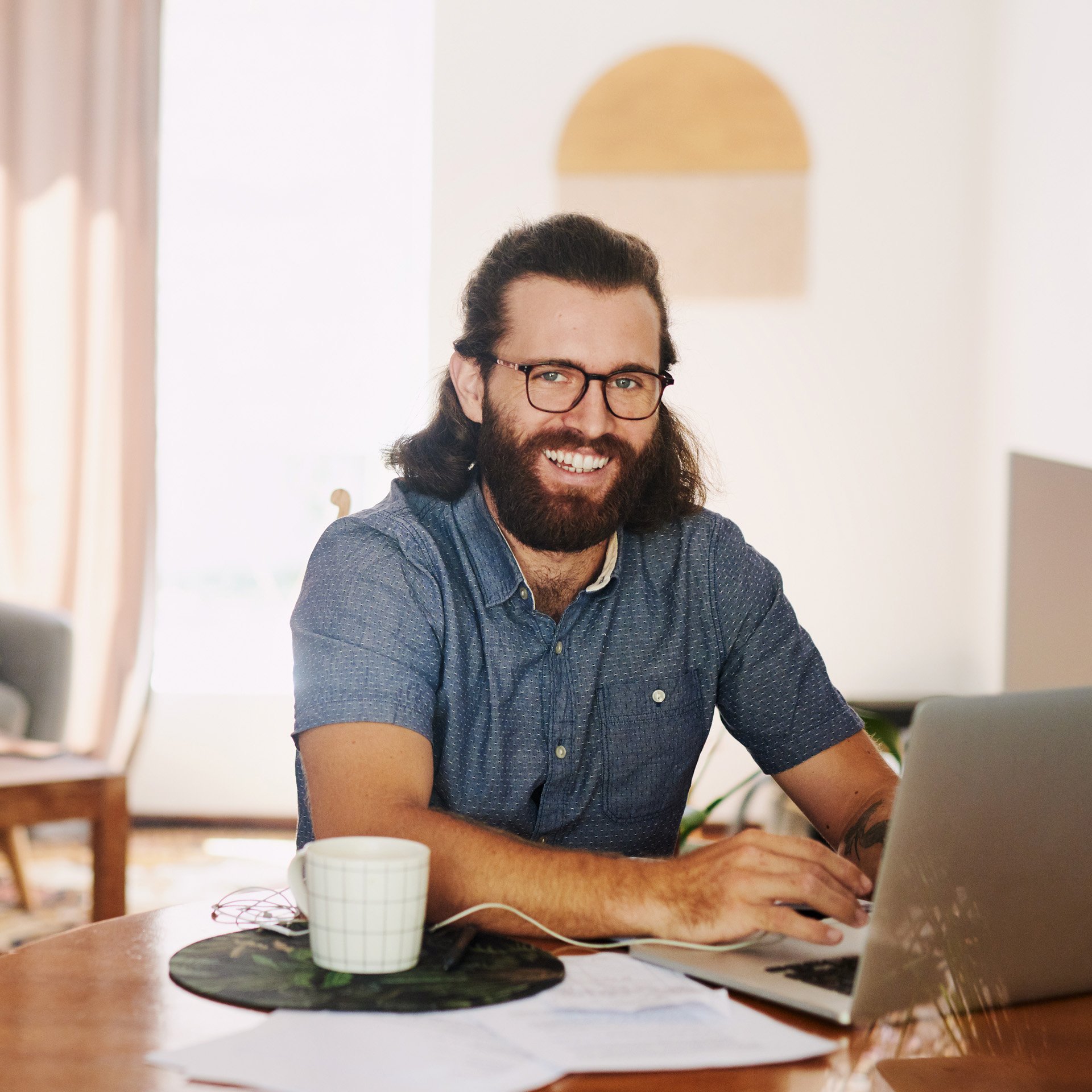 Main in short-sleeve blue shirt smiles at camera while studying online from a laptop at home