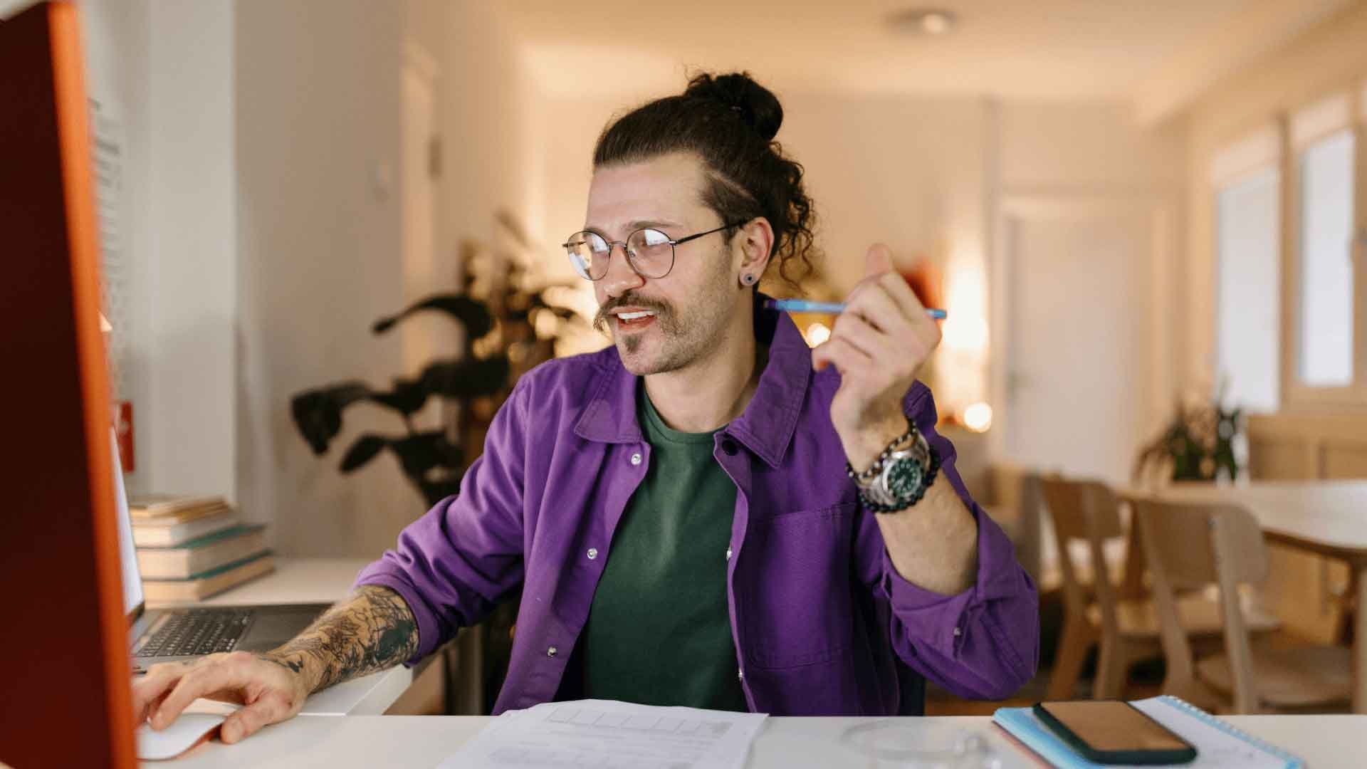 A man in glasses and a purple shirtsitting at a desk, holding a pen and looking at a laptop while studying a degree online.