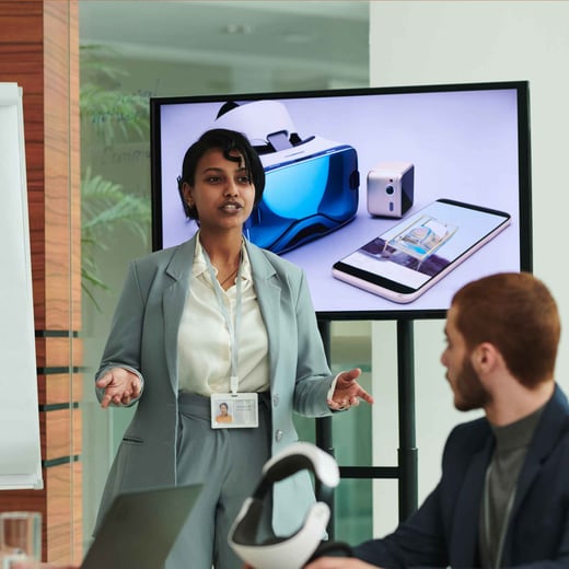 A business woman in a suit presenting in a meeting room, gesturing towards a screen while speaking to colleagues.