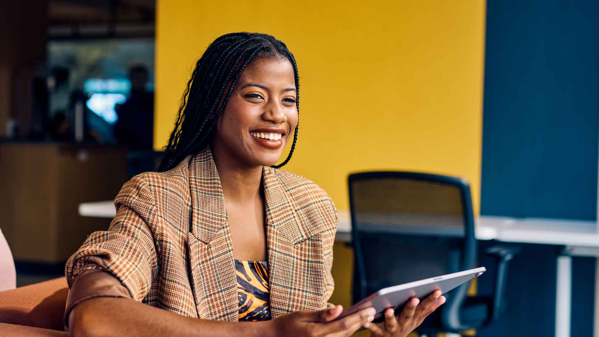 A businesswoman in brown blazer smiling while using a tablet in a modern workspace.