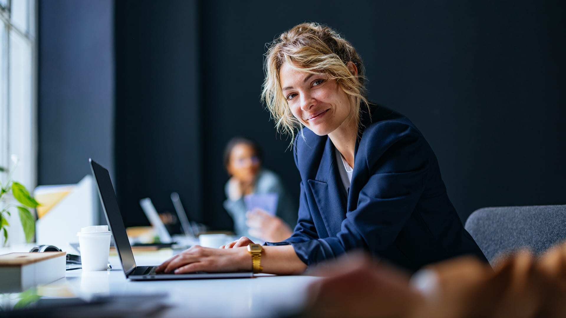 Professional woman in blue blazer sits at laptop and smiles at camera.