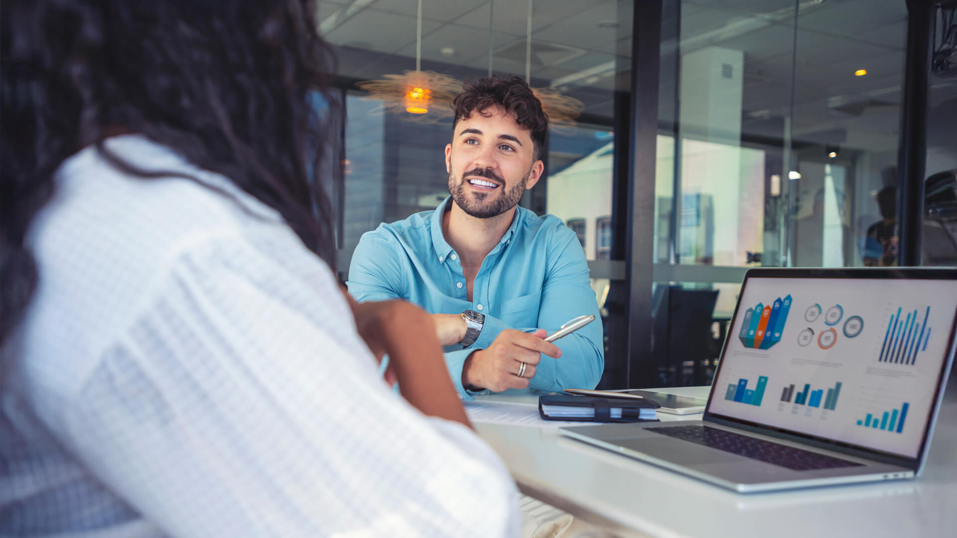 A man in a blue shirt talking with a colleague, discussing data reports on a laptop.