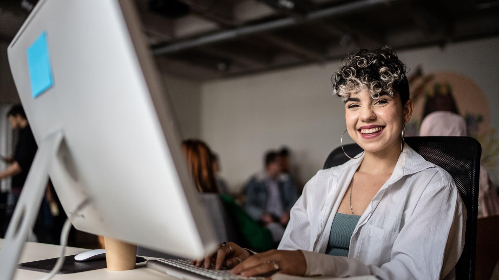 A smiling woman in white shirt working at a desk with a laptop and external monitor.