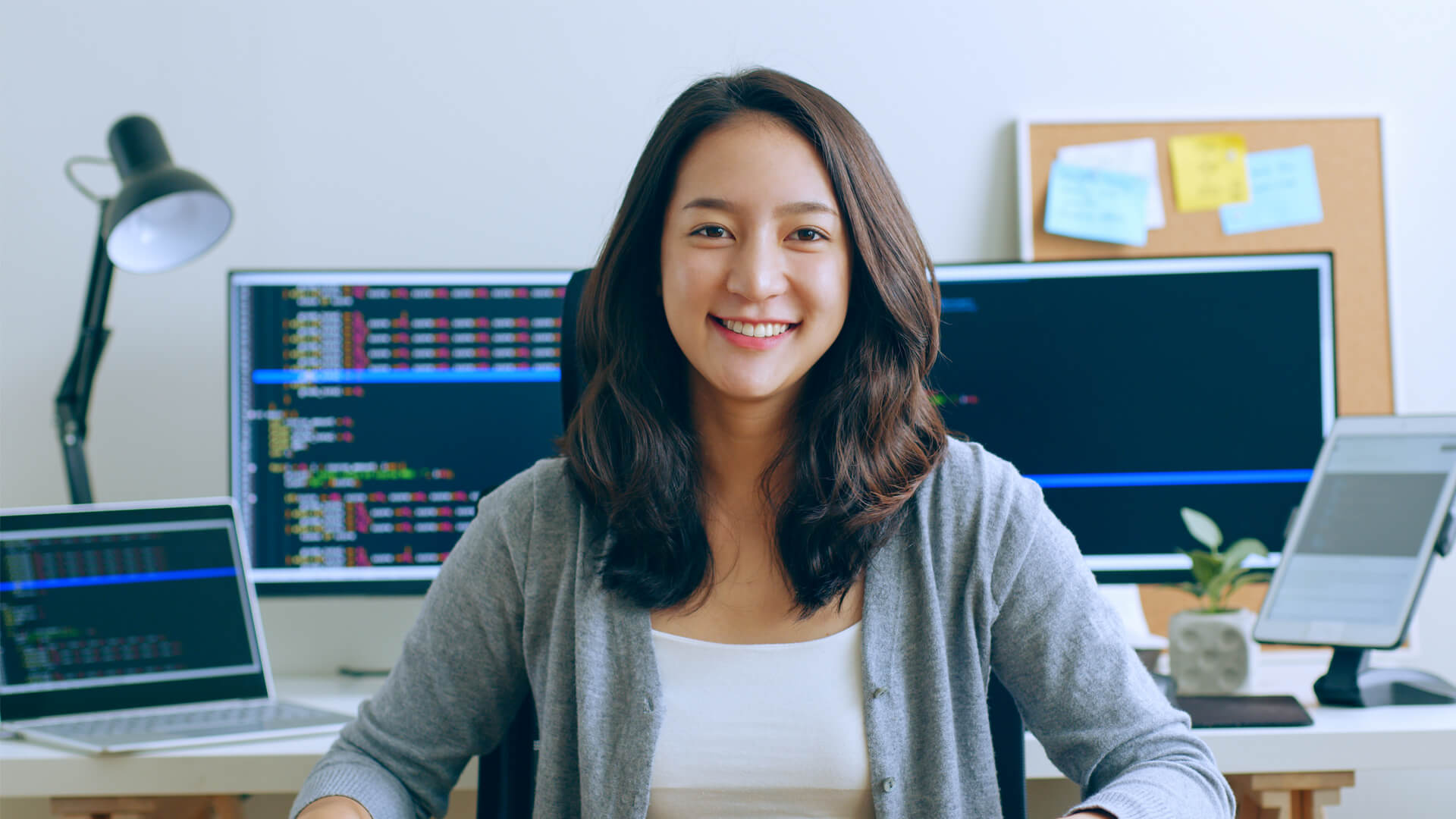 Smiling young professional sits at home desk with programming on computer screens behind her.