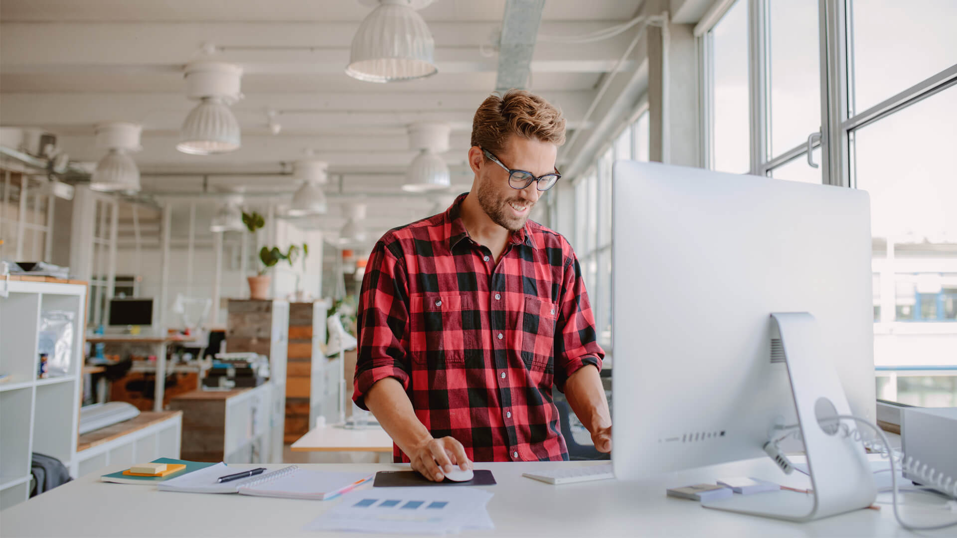 Man in red check shirt stands in modern office, working at computer at standing desk.
