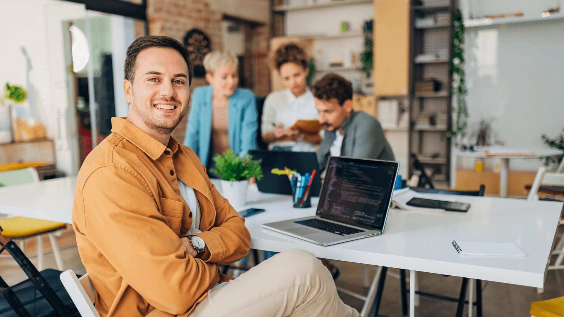 Man in yellow shirt sits at laptop that shows programming on screen, in front of colleagues