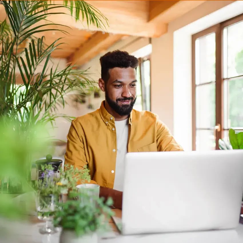 Man in yellow shirt surrounded by plants studying an online MBA from laptop