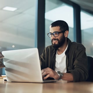 A man wearing glasses sat in an office, using the laptop in front of him to research what an MBA is