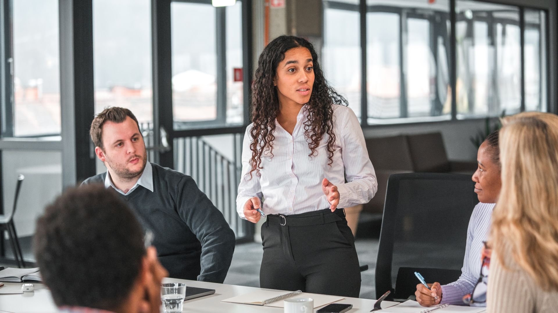 Woman standing up at a table, giving a talk to others in a meeting
