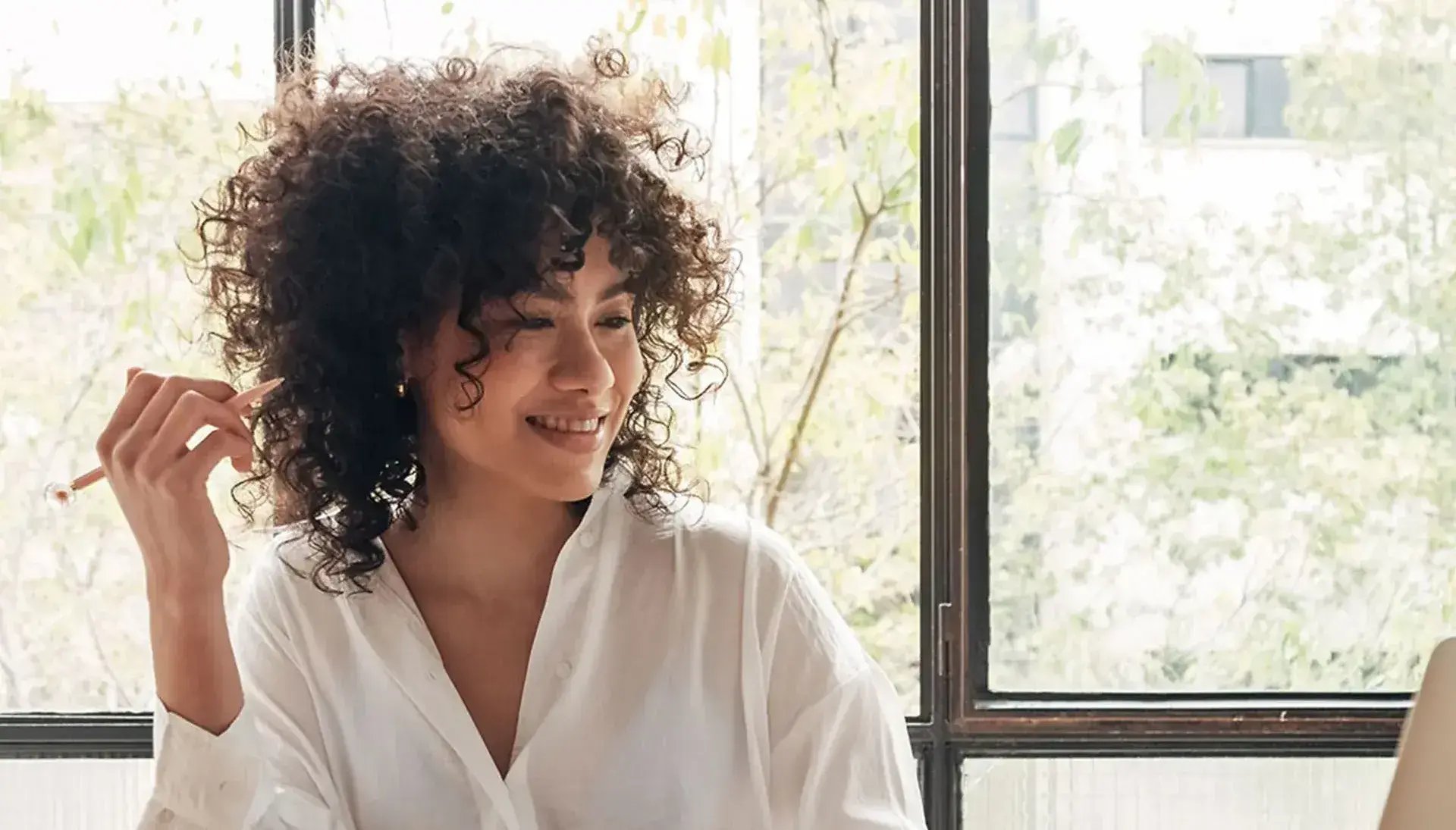 Young woman with curly hair holding a pen and looking at her laptop. On the table there is an open notebook and a mug.
