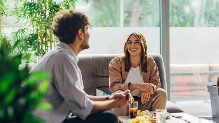 Two people sitting at a table by a large window, talking and smiling.