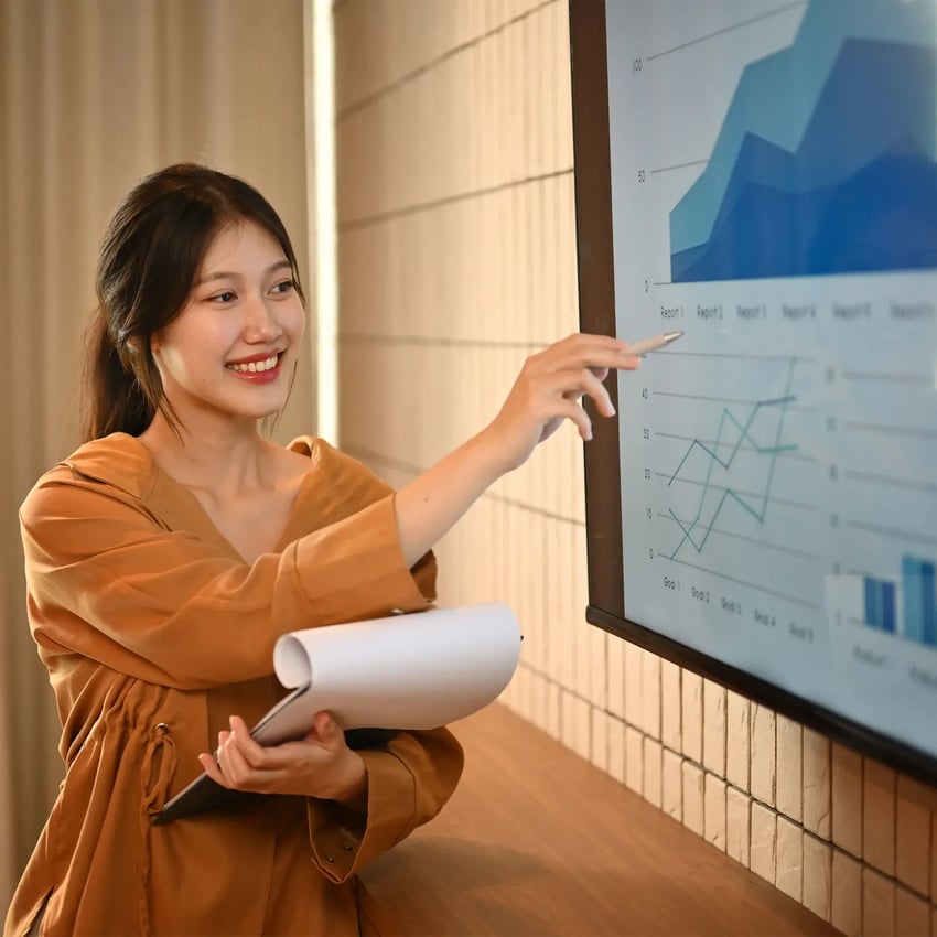 A young woman presenting and pointing at the screen with various graphs.