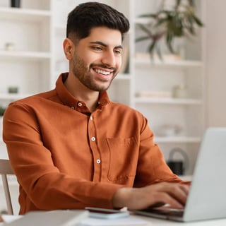 Young smiling man wearing an orange shirt, typing on his laptop.