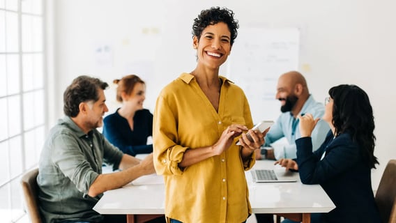 A woman standing with her phone and smiling. In the background there is a group of people sitting around a table and smiling.