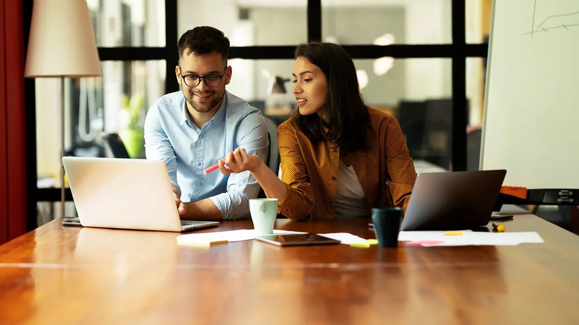 A man and a women sitting at a desk with laptops, mugs and papers.