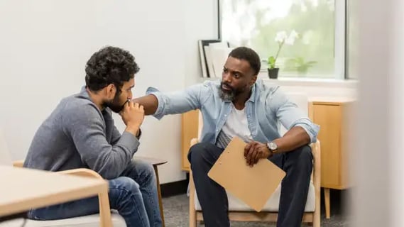 Male therapist in blue shirt has hand on shoulder of man seeking therapy.