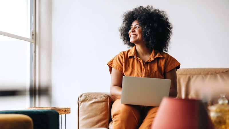 Woman in brown denim jumpsuit looks out of window with laptop on her lap
