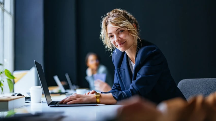 A confident mortgage adviser with a warm smile sitting at a table with a laptop, portraying positivity in her workplace environment.