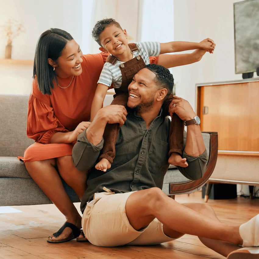 A joyful family moment at home, with a smiling woman embracing a little boy while he sits on a man's shoulders, celebrating his insurance certification.