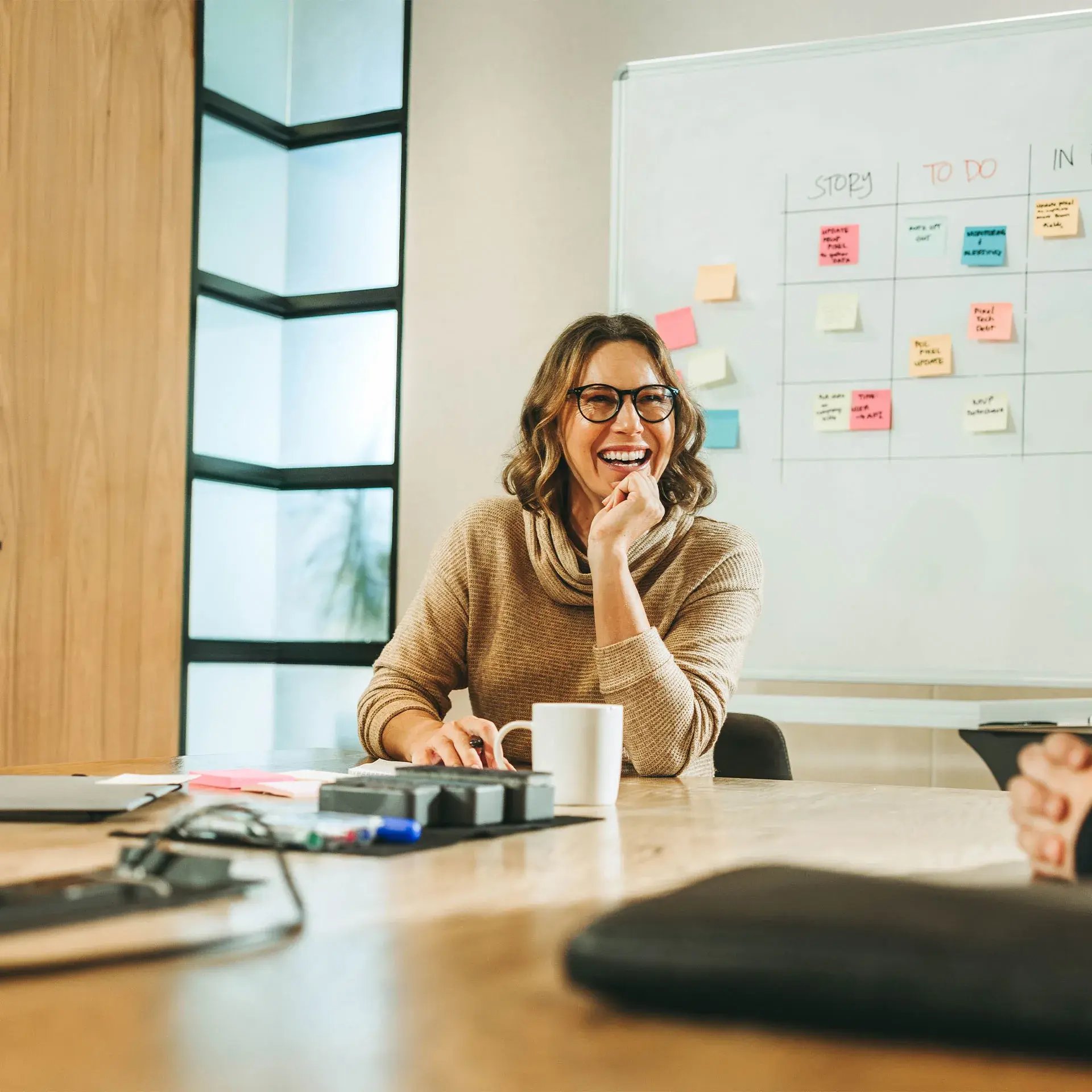 A woman wearing glasses is sitting at a table with a mug, holding a pen and smiling.