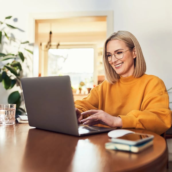 Young woman smiling, wearing glasses and an orange swear, typing on her laptop.