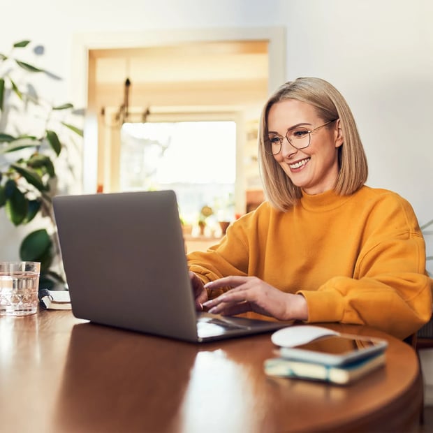 Young woman smiling, wearing glasses and an orange swear, typing on her laptop.