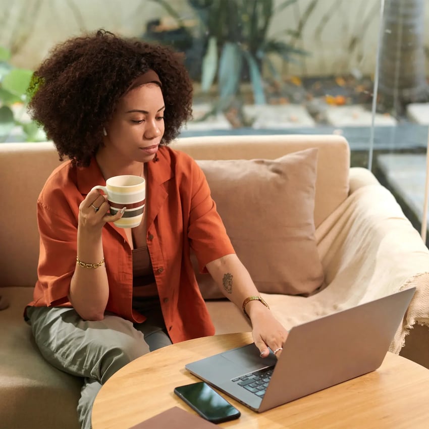 Girl wearing an orange shirt and holding a cup searches for Pearson VUE contact number uk on her laptop.