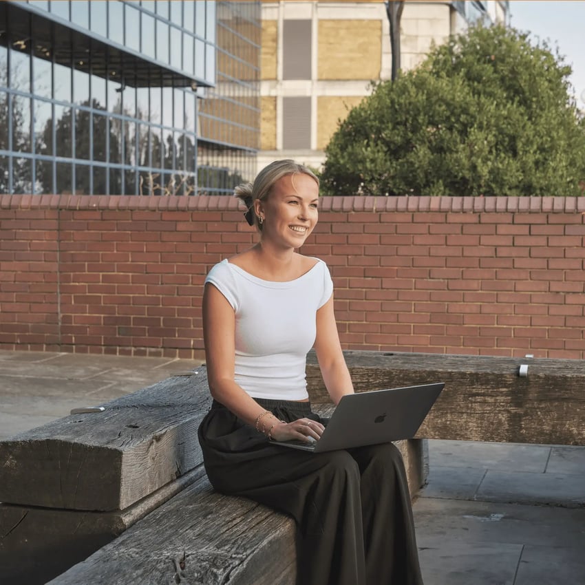 Smiling female student sits outside on laptop preparing for online assessment.